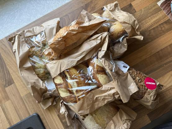 Eine Vielzahl von Backwaren, die in braunes Papier und Plastik eingewickelt sind, darunter Croissants und Brote, die auf einer Holzoberfläche angeordnet sind.
