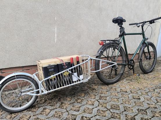 Ein Fahrrad mit Anhänger. Darauf ein großer Karton, zwei Öldosen und zwei Weinflaschen