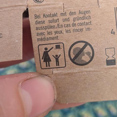 Foto eines Ausschnitts des Kartons, in dem das Shampoo war, mit Piktogrammen. Ein Piktogramm zeigt eine große und eine kleine Person. Die große streckt den Arm aus in Richtung eines Rechtecks mit Strich darunter, die kleine Person kommt mit ausgestreckten Armen nicht daran. Das Rechteck mit dem Strich könnte ein Regal sein, sieht aber auch aus wie ein überdimensionierter Zylinderhut. Daneben ist ein Kreis mit einem Strich durch. Dieser streicht den Kopf einer Person durch, die etwas trinkt.