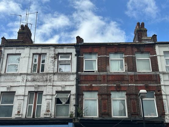 the top two stories of two brick row houses in a row; the left one is painted a (slightly peeling) white and, at some point, has its sash windows replaced with hinged ones, and the one on the right is unpainted, revealing slightly grimy tilework, suggesting a Victorian origin. The sky above is blue with white fluffy clouds.