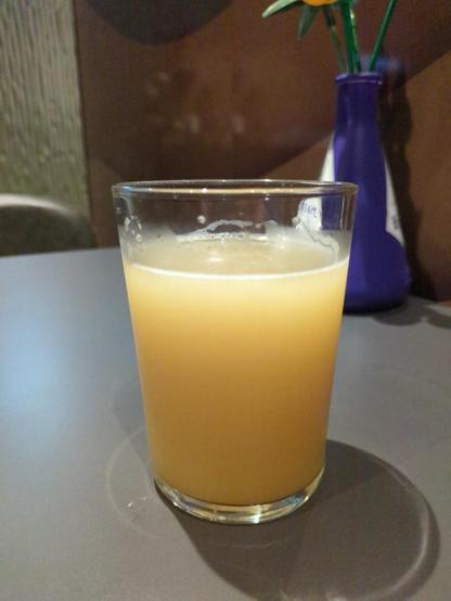 A glass of "Saturated" but the Feral Brewing Company. The glass is on a dark grey table against a brown wall. There is a conical purple vase behind the glass. The beer is opaque orange/brown colour. Due to the overhead lights there is a vertical gradient with it looking light yellow at the top. There is little to no head on the beer.