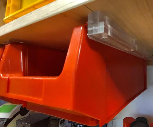 A red bin mounted below a shelf with a rail made from a transparent material.