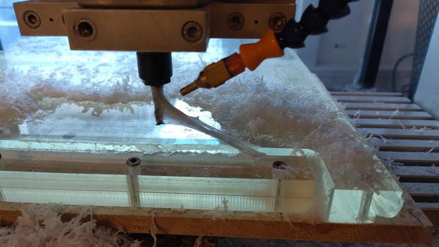Photograph of a small CNC mill milling a large piece of clear plastic. There are a lot of chips and a large blob of white goo sticks to the endmill looking a lot like spiderwebs.