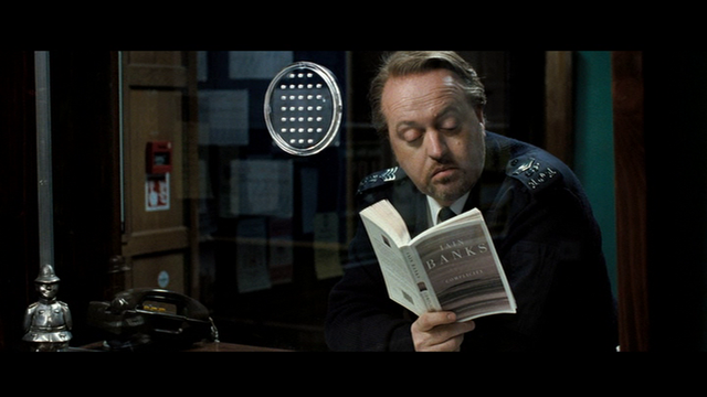 a still from Hot Fuzz showing Bill Bailey as one of the Sergaents Turner twins on duty reading Complicity by Iain Banks, roughly 12m37s into the film