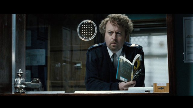 a still from Hot Fuzz showing Bill Bailey as the other of the Sergaents Turner twins on duty reading State of the Art by Iain Banks, roughly 42m39s into the film