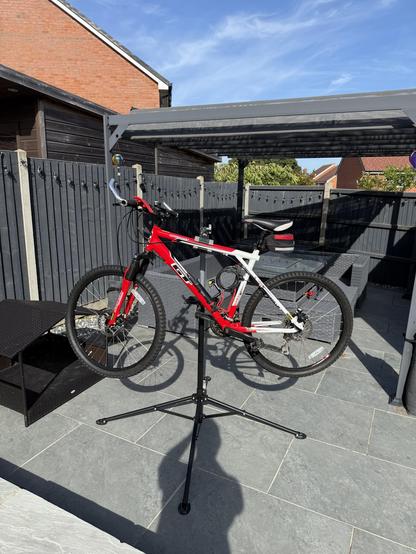 A red and white mountain bike on a black stand in a patio area with a wooden fence and a pergola.