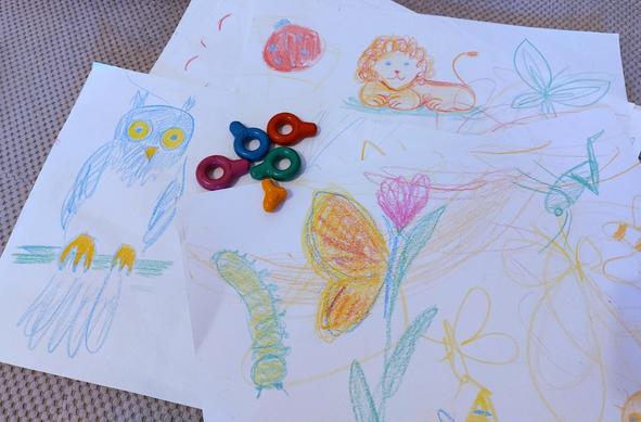 Large sheets of paper filled with simple, bright crayon drawings of animals (owl, lion, ladybug, caterpillar, butterflies, grasshopper) and random scribbled lines. There are 5 ergonomic crayons on the paper (blue, green, red, orange, pink). The yellow one is missing because its owner loves to run around the house holding it in hand.