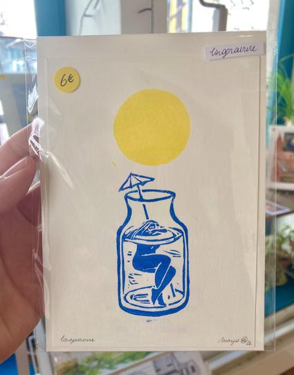 A photo of a linocut I am holding, sized A6. It is printed in 2 colours, blue and yellow. It shows a yellow, round sun and a little figurine in a glass vase swimming with a parasol printed in blue.