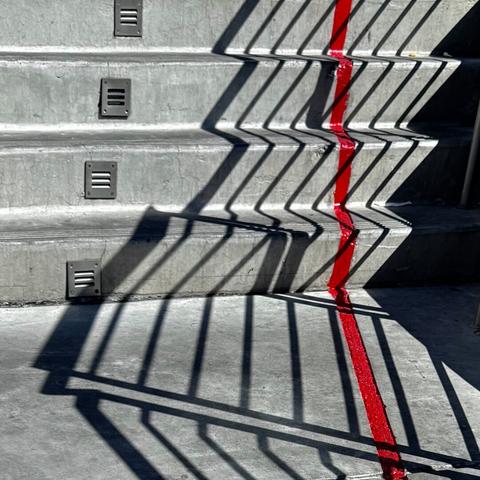 Railing shadows on a set of exterior concrete stairs. There is a single red stripe running up the right side of the stairs.