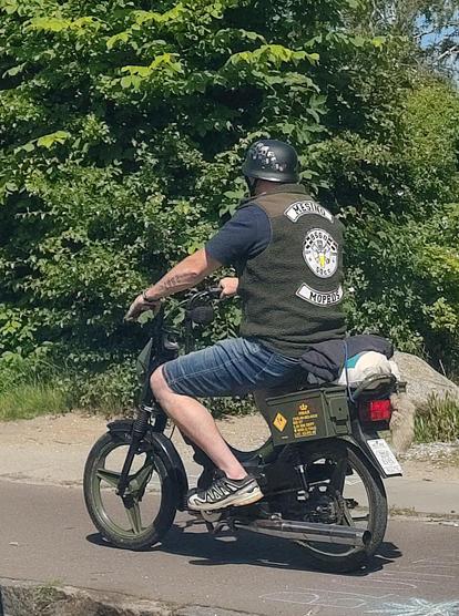 Man in his 40s riding an old 50cc moped (Puch Maxi perhaps?), dressed in a club vest and wearibg a classic moped helmet. A fox' tail is visible on the back of the moped.