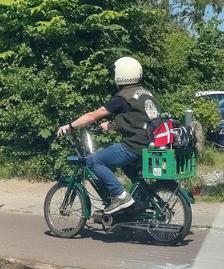 Man in his 40s riding an old 50cc moped (Puch Maxi perhaps?), dressed in a club vest and wearibg a classic moped helmet. A classic plastic milk carton crate on the back and a waiving Danish flag attached.