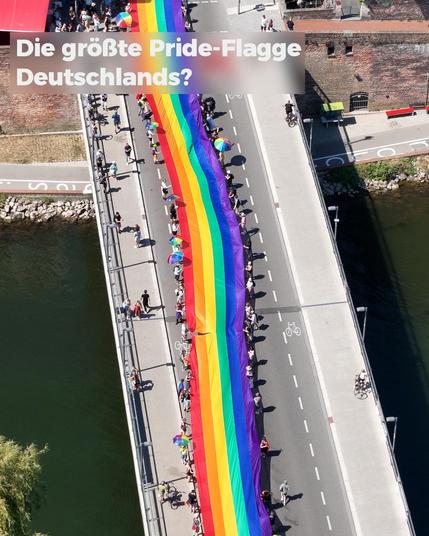 Luftaufnahme der Herdbrücke von oben, auf der viele Menschen rechts und links entlang einer vertikal durchs Bild laufenden Regenbogenflagge gehen und sie über die Brücke tragen