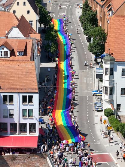 Luftaufnahme der gesamten Regenbogenflagge, die nach der Herdbrücke von vielen Menschen rechts und links der Flagge die Donaustraße im Bild vertikal nach oben getragen wird.