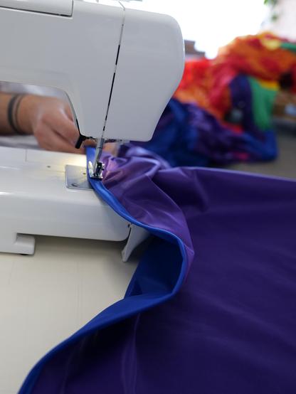 In der Mitte eine weiße Nähmaschine, in der der Saum des violetten Streifens der Flagge eingespannt ist. Im Hintergrund ist eine Hand zu erkennen, die den Saum in die Maschine einfädelt. Links im Hintergrund unscharf ein großer Haufen der bereits vernähten Regenbogenflagge.