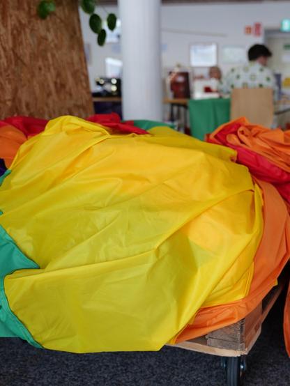 Im Vordergrund ist der bereits vernähte Teil der Regenbogenflagge auf einem Haufen liegend sichtbar, vor allem die gelben, orangenen und roten Felder. Im unscharfen Hintergrund sitzen mehrere Menschen an Tischen im Salon von Haus der Nachhaltigkeit und Temporärhaus und vernähen die Flagge weiter.
