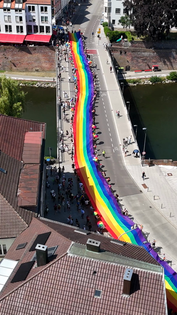 Hochkant-Video einer Regenbogenflagge, die von vielen Menschen rechts und links gehalten und über die Herdbrücke vom Neu-Ulmer zum Ulmer Ufer der Donau getragen wird.