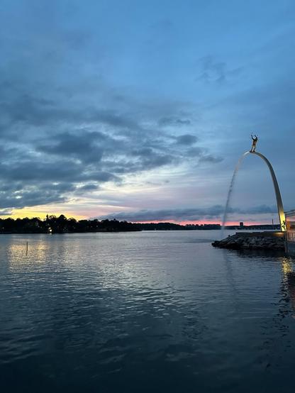 a view over water towards where the sun recently set. Clouds reflect in the choppy water. On the right stands a tall fountain shaped like a half-arch with a figure standing atop it.