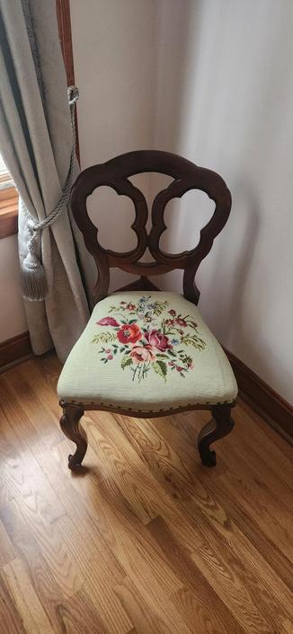 An ornate wooden chair, circa 1900, with an embroidered seat. It is in a corner with a heavy curtain on one revealing a sliver of window and high oak baseboards.
