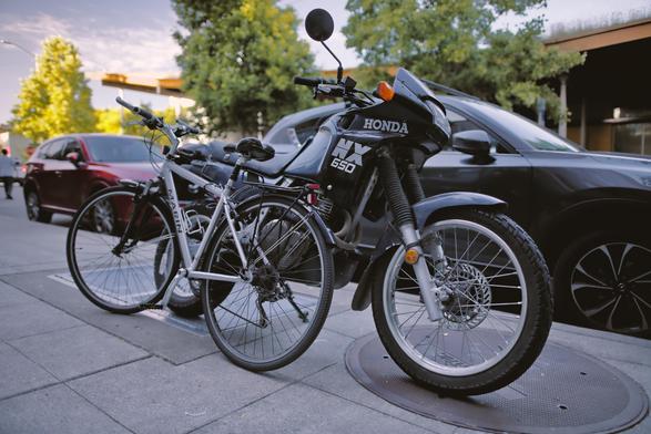 an urban sidewalk with cars and trees in the background. In the foreground are a modern Honda NX 650 and a Marlin flatbar bicycle are leaned against one another while facing opposite directions.