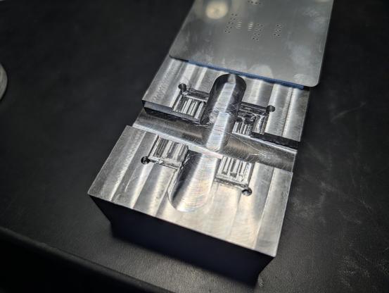 A small block of aluminum with features milled into its upper face. There is a deep X with 1/2" wide arms cutting entirely across the block, with a square sunk in, centered on the X.