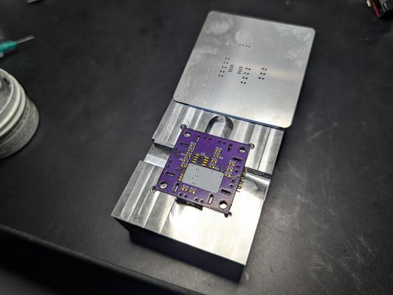The block with a square, purple PCB perfectly nestled into the milled out cavity. A stainless steel solder mask is folded back above it.