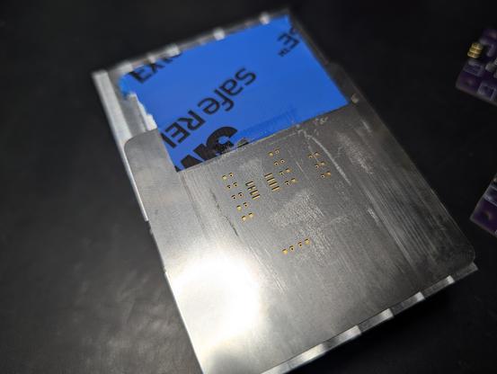 The solder mask folded down to cover the PCB, leaving only the solder pads visible though its holes. It has been taped to the aluminum block with some blue masking tape.