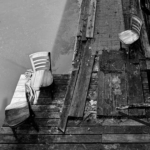 Black and white photo of two wooden chairs sunbathing on a deteriorating dock while another chair looks on from the shadows.