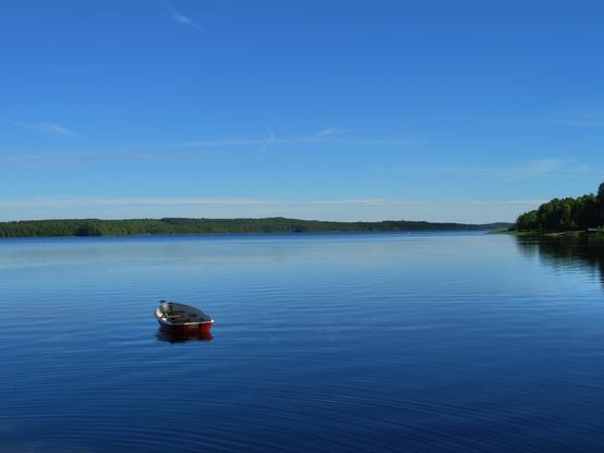 Photograph of a lake, with a red and white boat in the shot. Lake is still, sky is clear blue.