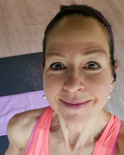 Unsere Kursleiterin für Yoga, Iris Schaub, im pinken Sporttop macht Selfie auf Yogamatte mit hellem Holzboden im Hintergrund.