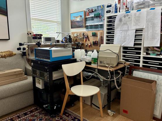 A workbench, rolling cart, and rolling table covered in electronics gear, inelegantly shoved into a corner.