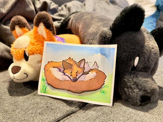 A photo of a watercolor painting featuring a tiny fox worm surrounded by two fox worm plushies
