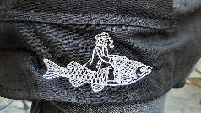 A lindrawing in white on black cloth, showing a fisherman wearing a caprains hat and a kaban coat, riding a big fish