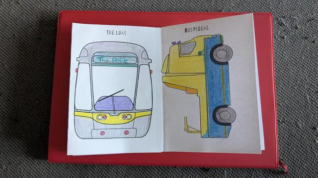 Irish transport icons zine open on a red diary sitting on a grey cushion. The zine is open on the LUAS and Buspidéal, coloured in with pencil