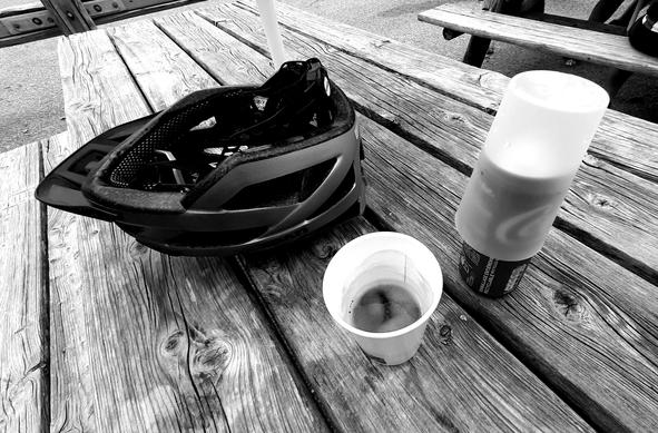 Picture of my helmet, a Coffee in a paper cup and a can of Coke on a rough wooden tabletop