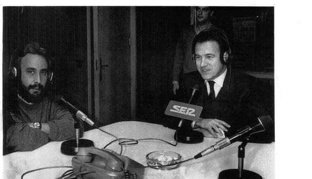 Imagen antigua (años ochenta) de un estudio de radio con dos personas, entrevistador y entrevistado,  en una mesa con varios micrófonos, un teléfono y un cenicero. Al fondo, casi oculta, una tercera persona.