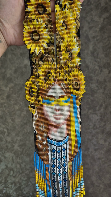 ​A close-up of a stunning, handmade traditional Ukrainian beaded necklace known as a "gerdan." The necklace is finely woven bead-by-bead into a detailed tapestry. The central design features a beautiful portrait of a young Ukrainian woman wearing a massive, vibrant crown made of bright yellow sunflowers that extend up the straps of the necklace. Her long brown hair flows down, with a striking blue and yellow flag-colored strand woven into it. Her gaze is symbolically shielded by stylized blue and yellow glasses or bands, representing resilience. She wears a traditional embroidered blouse (vyshyvanka) pattern in blue, white, and black. The bottom of the gerdan finishes with intricate, long beaded fringes in shades of sky blue, yellow, and white that elegantly hang down.