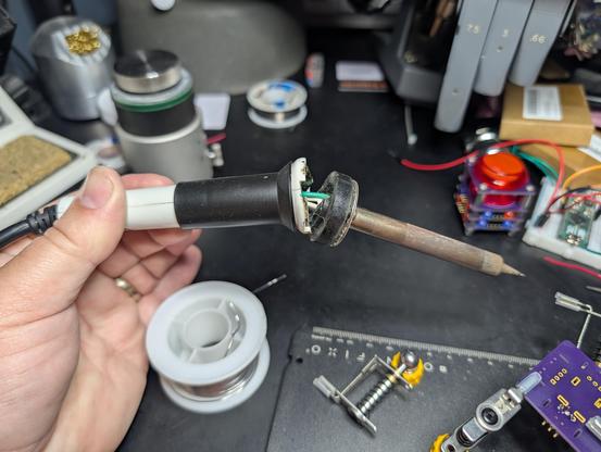 A soldering iron being held above a surprisingly lightly cluttered electronics bench. The plastic on the core of the handle has broken, exposing internal wiring and letting the iron droop sadly.