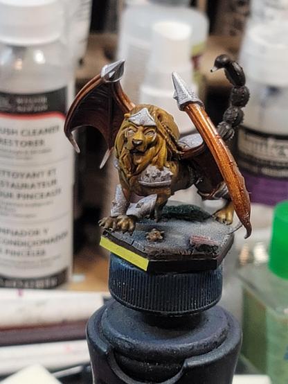 A battletech hex base, with yellow 'front' showing.  Atop it, the terrain is damaged concrete, a bit of downed rusty sheet metal fencing, a torn up teddy bear, and some areas where grass has fought its way through the concrete.  Perched atop it, standing over the teddy bear as if to protect it, is a bizarre and decidedly non-mechanical creature with the body of a lion, wings like a bat, and the tail of a scorpion, with armored bracers showing on its forelegs, shoulders, and forehead. The fur has been done in a nice golden brown, the nose in dark brown, the wings in a base rusty red, and the tail in a brown so dark it is nearly black.