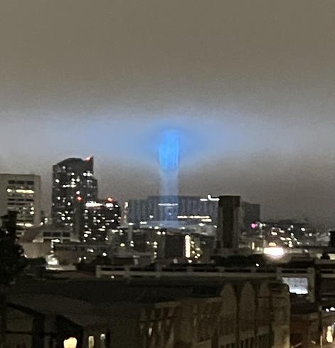 A nighttime cityscape featuring various skyscrapers, with a prominent blue light beam shooting straight upwards into the fog.