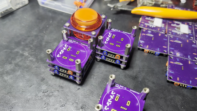 Four stacks of purple PCBs. One has a large red button on the top, while the others are incomplete. Three are sitting next to each other in a V. A finger pushed the 4th into the notch, and magnets snap it into place.