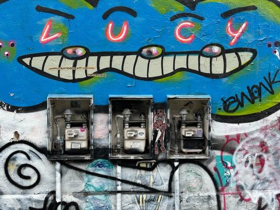 three gas meters in enclosures against a graffiti-covered wall, the bottom of which is mostly white (with some colours showing through). Above the meters is a blobby blue and green piece shaped sort of like three faces melted together, with the eyes replaced with the letters LUCY.