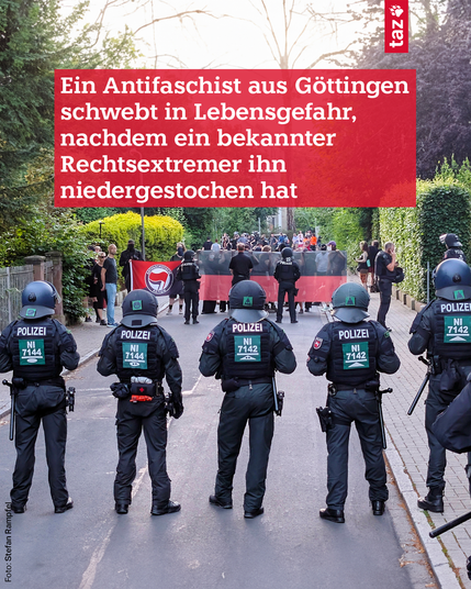 „Ein Antifaschist aus Göttingen schwebt in Lebensgefahr, nachdem ein bekannter Rechtsextremist ihn niedergestochen hat“. Foto: Rückansicht von fünf Polizistinnen und Polizisten in schwarzer Einsatzmontur und Helmen, auf deren Rücken „POLIZEI“ und „NI 7142“ oder „NI 7144“ steht. Sie stehen auf einer Straße und blicken auf eine Gruppe von Menschen, darunter Personen mit einem roten Banner und einer transparenten Barriere. Im Hintergrund sind Bäume und Sträucher sichtbar.