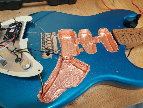 A blue electric guitar's body which has been opened up. The cavities have been covered with copper tape, the pickguard already has a metal shield.