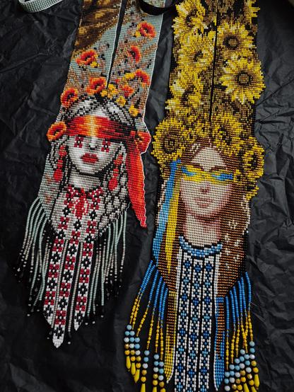 A top-down photograph of two intricate handcrafted beaded gerdan necklaces. The left one depicts a woman with red poppy motifs and a blindfolded face, flanked by geometric patterns. The right one features a woman with a sunflower crown and blue and yellow elements over her eyes. Both pieces have beaded fringe and lie on a textured black paper surface.