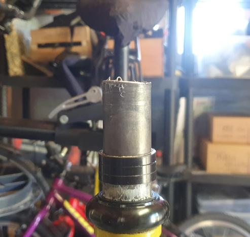 Image d'un tube de fourche de vélo au premier plan, vu de devant. On distingue le cadre jaune en bas.
Flou, à l'arrière plan, un bazar de garage/atelier.
Le tube semble coupé de biais.