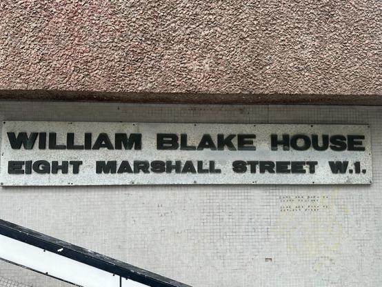 a sign on a tiled wall of a modernist building, reading “WILLIAM BLAKE HOUSE, EIGHT MARSHALL STREET W.1.”. Below the right side of it, someone has written something on the grid of tiles
