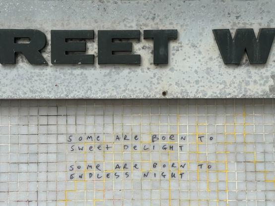 the tiled wall beneath the sign. A graffito inscribed in the grid of small white tiles reads “some are born to sweet delight, some are born to endless night”