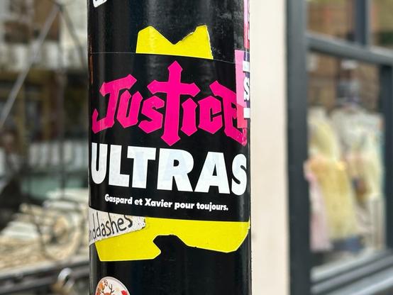 a sticker on a post reading “JusTicE ULTRAS” (with the logo of the French EDM band Justice) and below that, “Gaspard et Xavier pour toujours”
