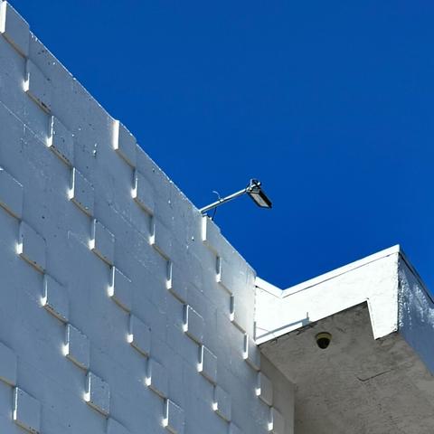 White building with a decorative wall with raised blocks where it's mostly cast in shadows aside from little squares of light near the bottom of each block. Beyond is a bright blue sky.