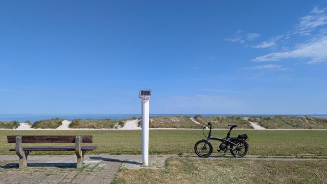 Auf der Deichkrone steht rechts ein Tern Faltbike, mittig ist eine Notrufsäule, links daneben eine Bank. 
Dahinter ist der abfallende Deich und die Dünen. 
Im Hintergrund ein Streifen Ostsee und der blaue Himmel.